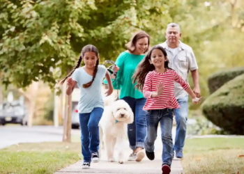 A joyful family with two young girls and a fluffy white dog walk on a tree-lined sidewalk. The children run ahead, conveying excitement and happiness.