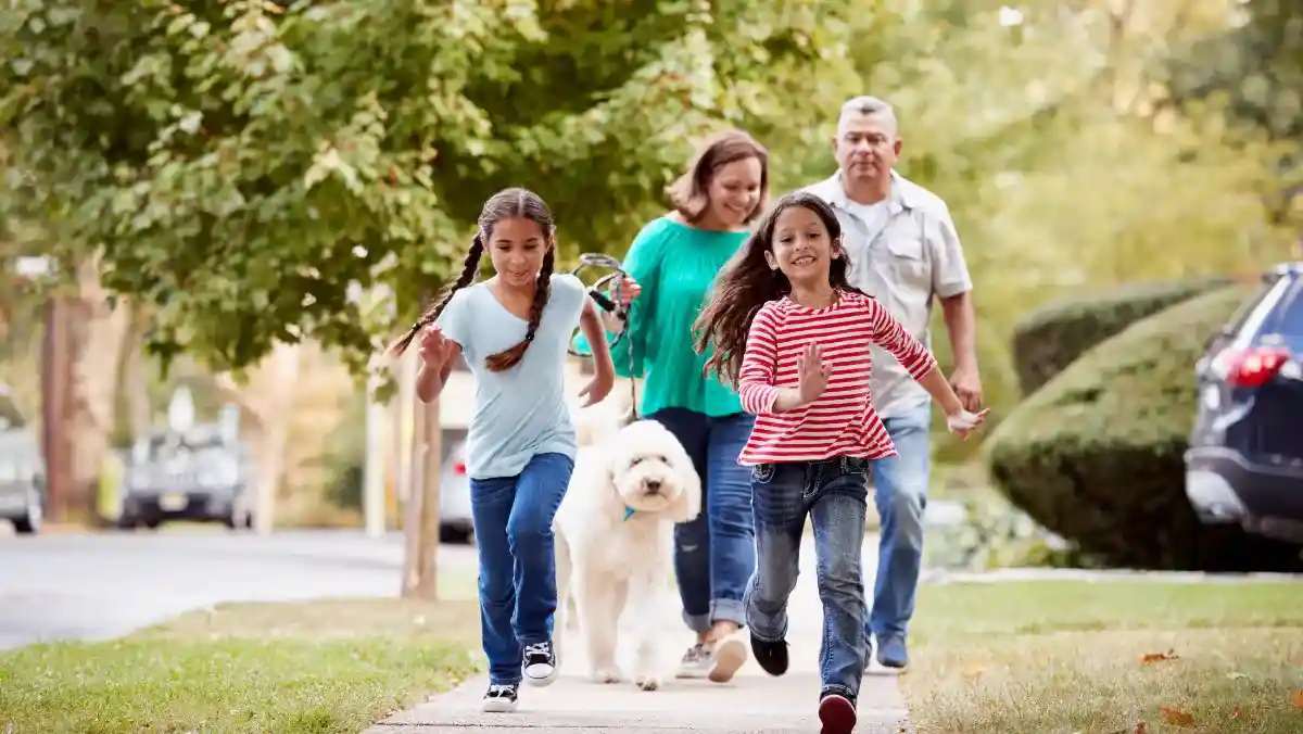 A joyful family with two young girls and a fluffy white dog walk on a tree-lined sidewalk. The children run ahead, conveying excitement and happiness.