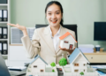 Asian businesswoman holding a house model in front of a laptop, symbolizing real estate or property investment.