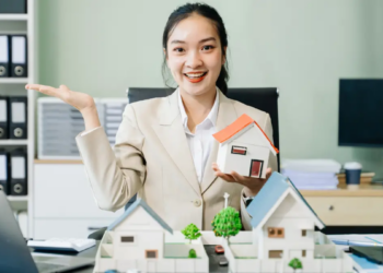Asian businesswoman holding a house model in front of a laptop, symbolizing real estate or property investment.