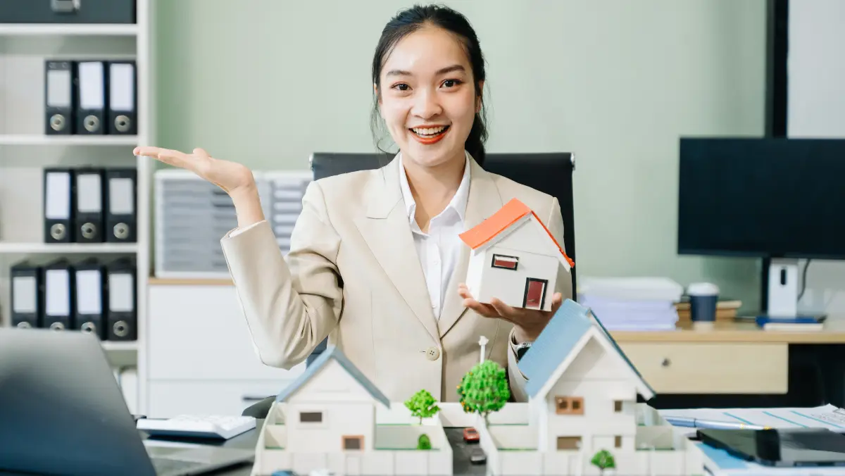 Asian businesswoman holding a house model in front of a laptop, symbolizing real estate or property investment.
