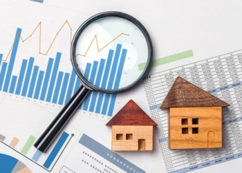 Magnifying glass on bar graph charts, two wooden houses on top of printed Excel sheets.