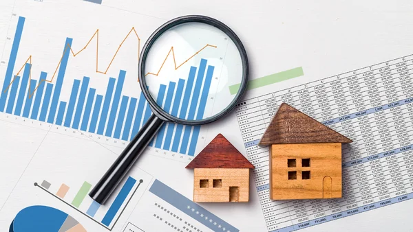 Magnifying glass on bar graph charts, two wooden houses on top of printed Excel sheets.