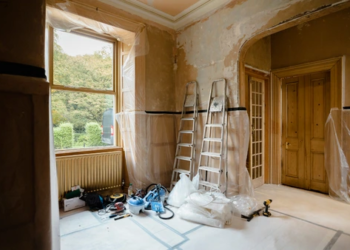 A room undergoing renovations, stripped to the plaster. Supplies and ladders sit in one corner beside a window.