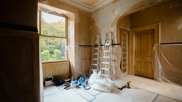 A room undergoing renovations, stripped to the plaster. Supplies and ladders sit in one corner beside a window.