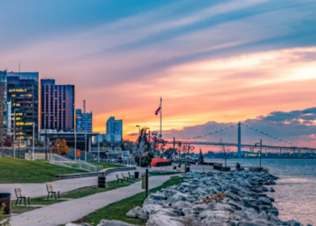 Windsor Ontario skyline, showing a bayfront walkway with tall buildings and a bridge in front of the sunset.