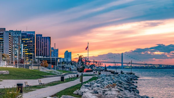 Windsor Ontario skyline, showing a bayfront walkway with tall buildings and a bridge in front of the sunset.