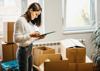 A woman looking at a clipboard in a room full of cardboard boxes.