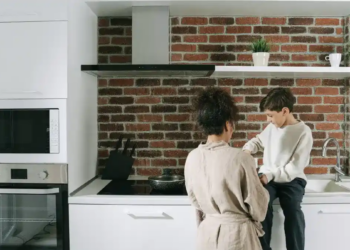 In a kitchen featuring a brick wall, two people stand side by side, smiling and interacting with each other.