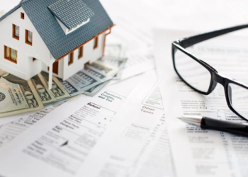 A small wooden house on top of some money and tax forms.