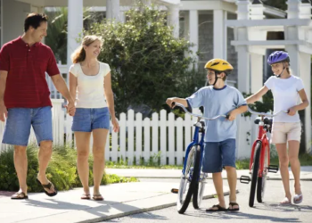 Two kids walking with bikes and their parents watching from the yard.