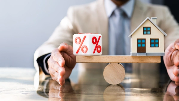 A wooden house and dice with a percent sign balance on a wooden beam.