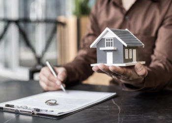 A person signs a document with one hand and holds a model home in the other