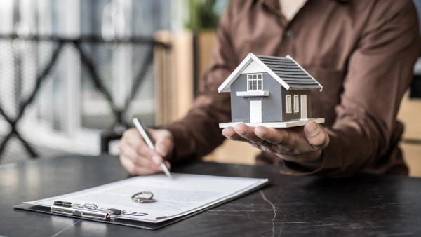 A person signs a document with one hand and holds a model home in the other