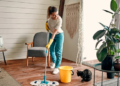 A woman using a mop and bucket to clean the floors with her cat on the ground.