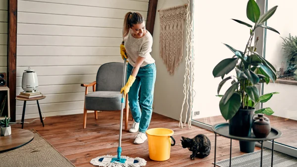 A woman using a mop and bucket to clean the floors with her cat on the ground.