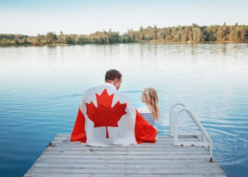 A father and daughter sit on a dock with a Canadian flag draped over their shoulders.