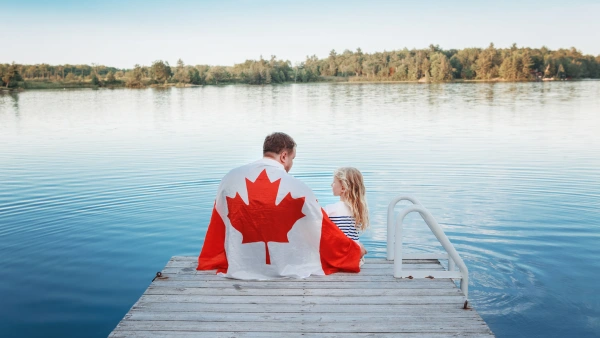 A father and daughter sit on a dock with a Canadian flag draped over their shoulders.
