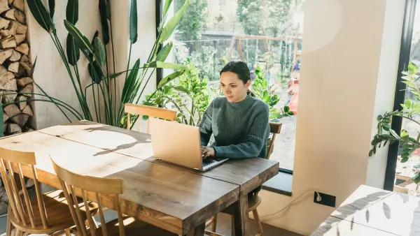 A woman who is sitting at a wooden desk, working on her laptop surrounded by green plants and a large window.