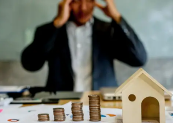 A person stressed out behind a small wooden house and coins beside it.