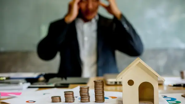 A person stressed out behind a small wooden house and coins beside it.