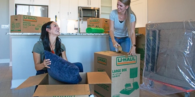 Two women laugh together as they work on packing items from their living room into U-Haul moving boxes to prepare for moving day