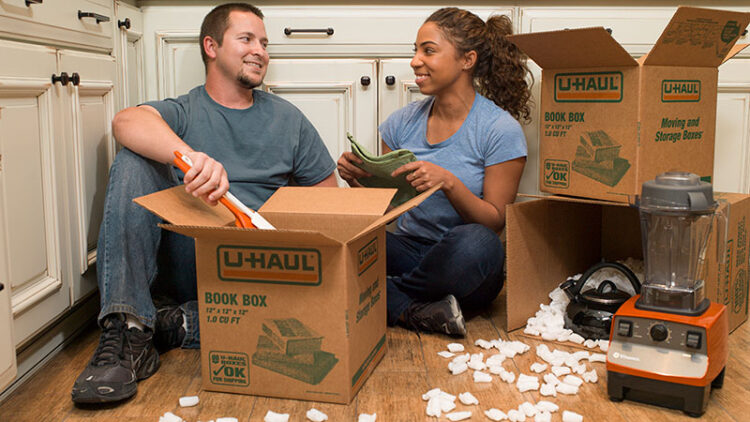 A couple sitting on the ground looks at each other as they pack their kitchen supplies using U-Haul packing peanuts