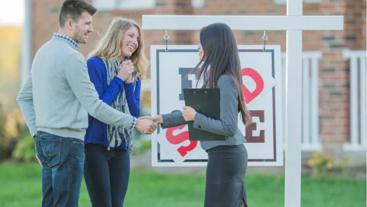 A real estate agent shaking hands with a man, while another woman stands next to him. All three are in front of a 'for sale' sign outside a house