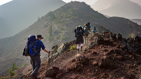 Three people carrying backpacks hike upward on a mountain, with scenic views of nature surrounding them.