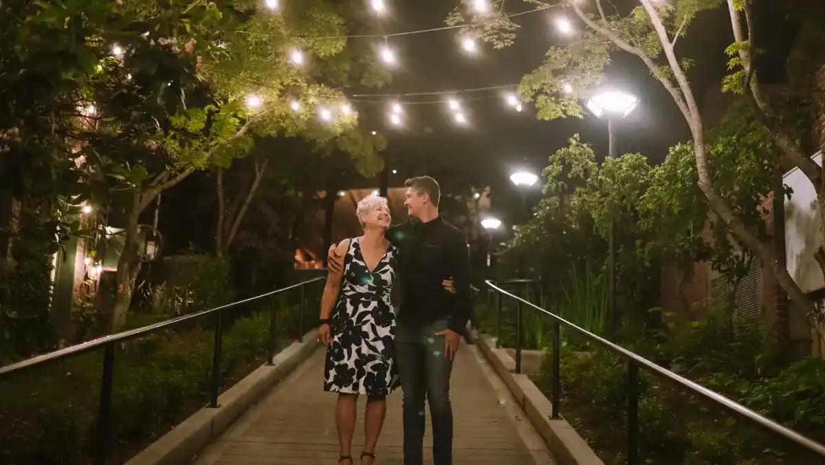 A couple walks hand in hand along a path illuminated by string lights.