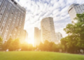 A city park featuring green grass and trees, with tall buildings rising in the background under a clear blue sky.