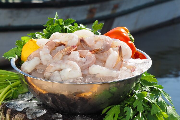 A bowl of shrimp and vegetables sits on a table in front of a boat.