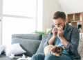 A man on a couch with a dog, using a tissue to blow his nose.