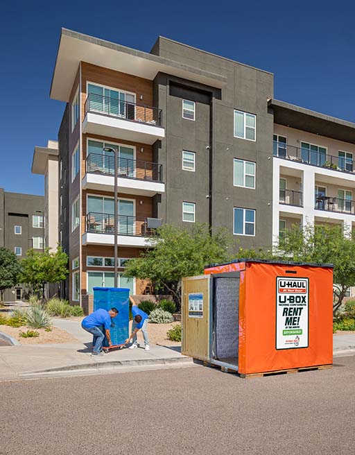 Two male Moving Help Service Providers carefully move a heavy piece of furniture on a furniture dolly while heading toward a U-Box storage container.