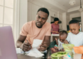 A dad examining his finances at the kitchen table while his child eats next to him.