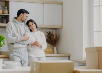 A couple stands in a ktichen smiling. Moving boxes are surrounding them.