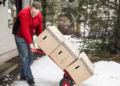 A man uses a dolly to carry moving boxes into his house while walking in the snow.