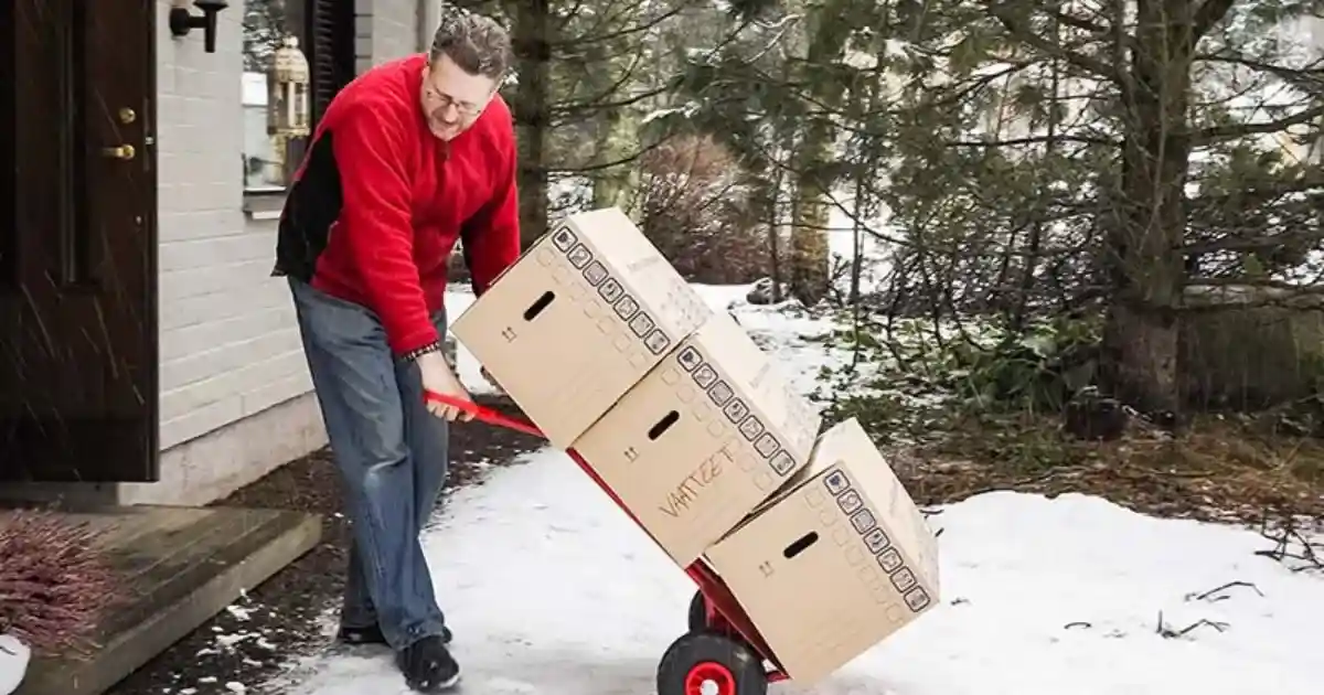 A man uses a dolly to carry moving boxes into his house while walking in the snow.