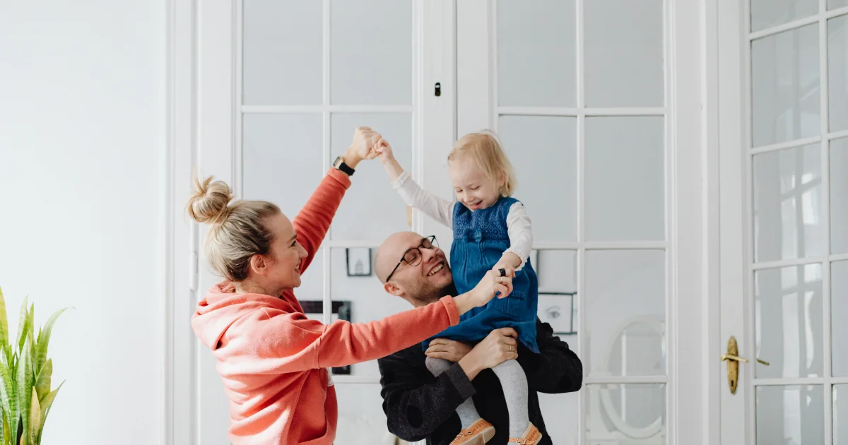 A dad holds a young daughter on his shoulders while a mother raises her hands in the air happily.