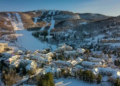 Aerial view of a snowy ski resort village nestled in a valley with winding roads. Snow-laden trees and ski slopes on the mountain create a serene winter scene.