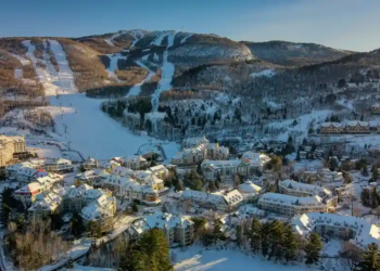 Aerial view of a snowy ski resort village nestled in a valley with winding roads. Snow-laden trees and ski slopes on the mountain create a serene winter scene.