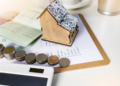 A small house model next to a stack of coins on a wooden desk, symbolizing real estate investment and savings.