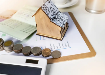 A small house model next to a stack of coins on a wooden desk, symbolizing real estate investment and savings.