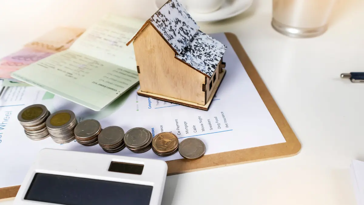 A small house model next to a stack of coins on a wooden desk, symbolizing real estate investment and savings.