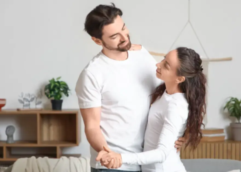 A married couple stands in a living room holding each other.