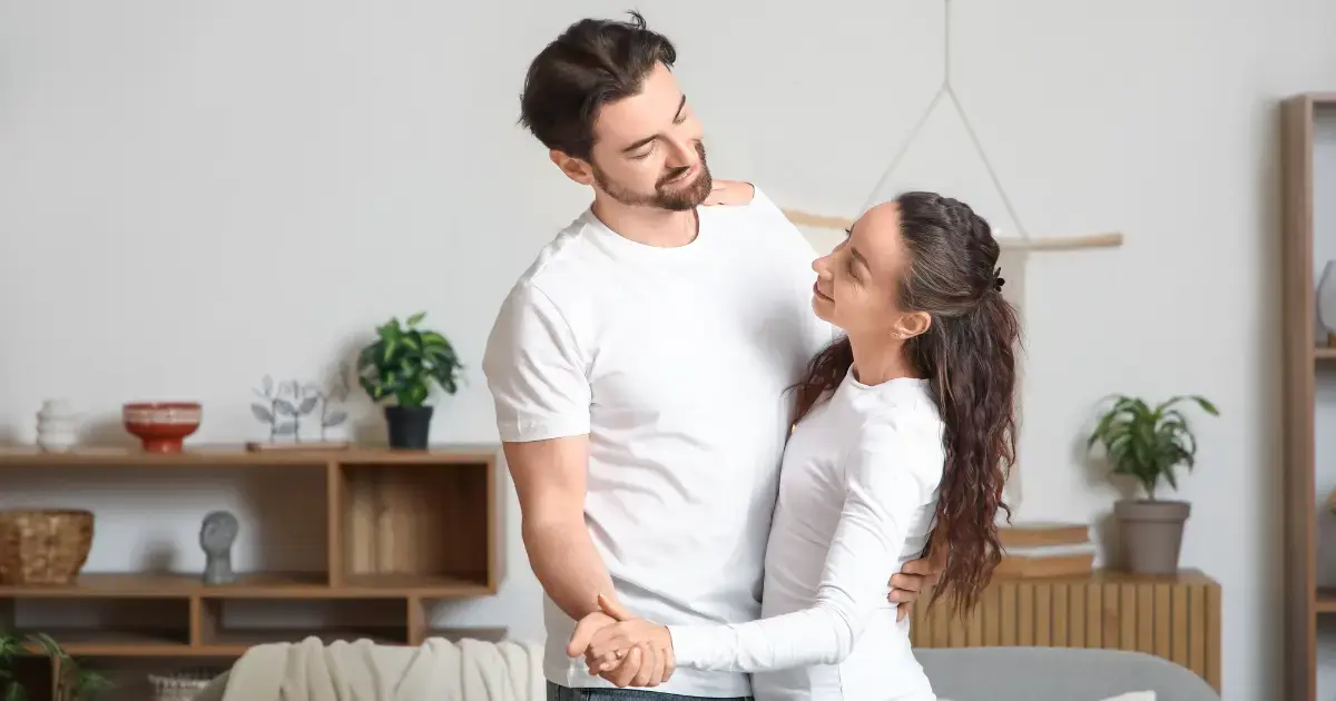 A married couple stands in a living room holding each other.