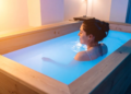 Woman relaxing in a wooden floatation tank filled with water, illuminated by soft blue light. The ambiance is peaceful and serene, conveying tranquility.