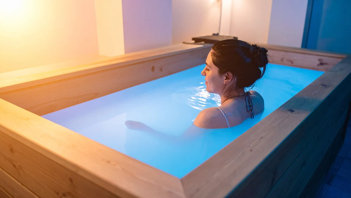 Woman relaxing in a wooden floatation tank filled with water, illuminated by soft blue light. The ambiance is peaceful and serene, conveying tranquility.