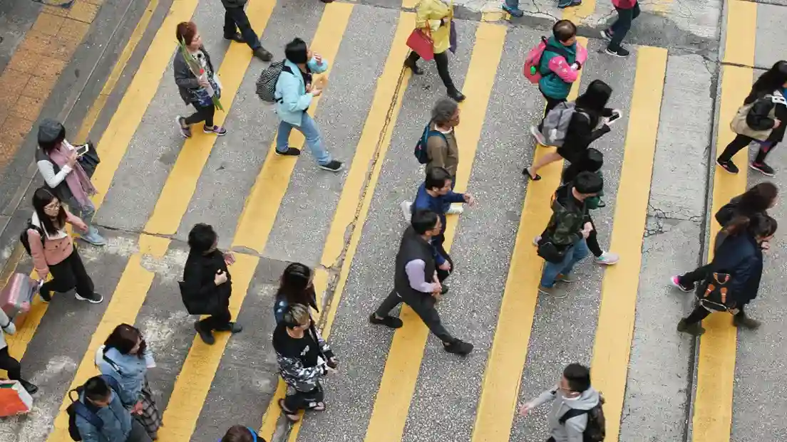 Aerial view of a busy city crosswalk with diverse people crossing in different directions. Yellow stripes highlight the lanes, and the scene feels dynamic.