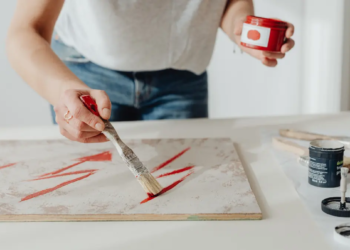 A person in a white shirt paints red brushstrokes on a canvas using a wooden brush. Various paint jars are scattered on a white table, creating an artistic and focused atmosphere.